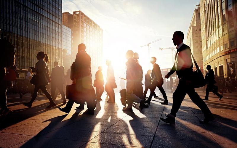 Group of pedestrians walking through a city street with sun shining from behind.